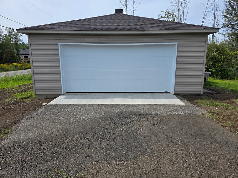 Dalle de béton finie devant la porte double d'un garage détaché, raccordée à l'entrée de gravier