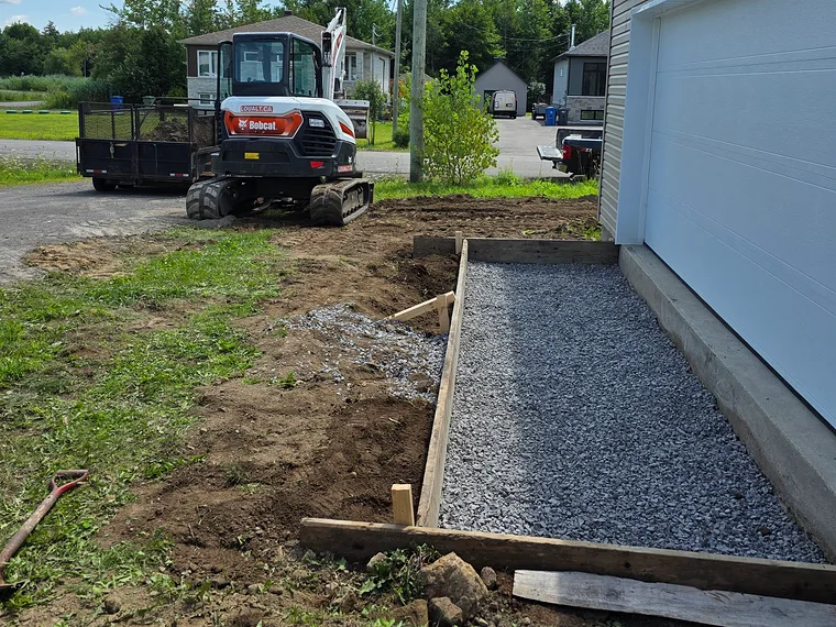 Avant : le lit de gravier et le coffrage de bois le long du garage, la mini-excavatrice de l'équipe stationnée derrière