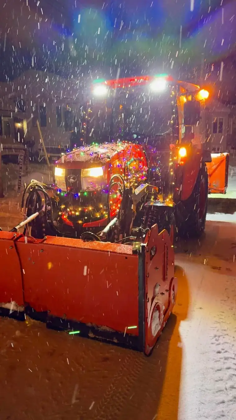 Tracteur de déneigement décoré de lumières de Noël, lame relevée, en pleine chute de neige la nuit